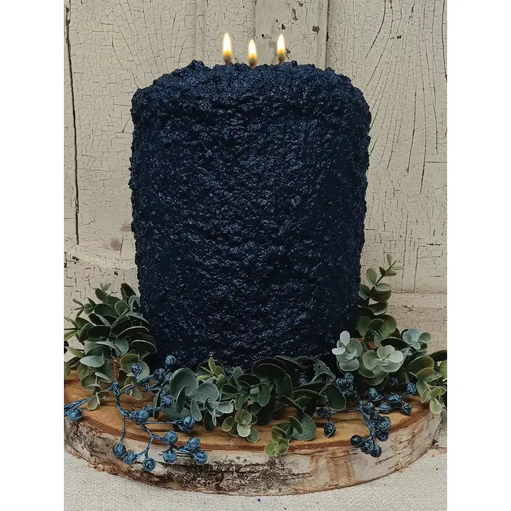 A large, dark blue candle with three lit candles sits on a wooden base, surrounded by greenery and blue flowers.