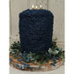 A large, dark blue candle with three lit candles sits on a wooden base, surrounded by greenery and blue flowers.