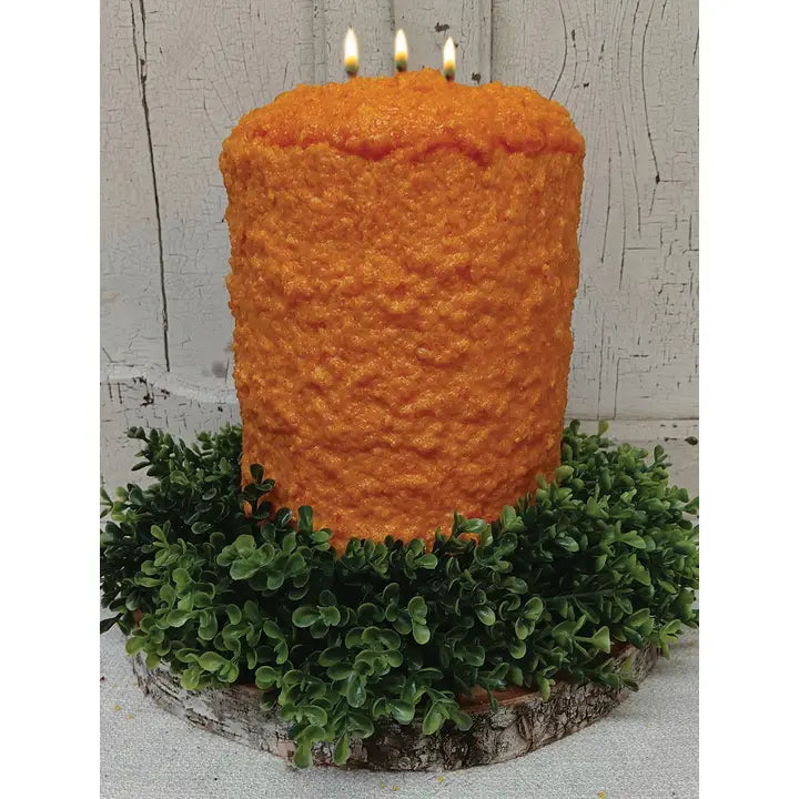 A large orange candle with three lit candles sits on a wreath of green foliage.