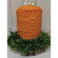 A large orange candle with three lit candles sits on a wreath of green foliage.