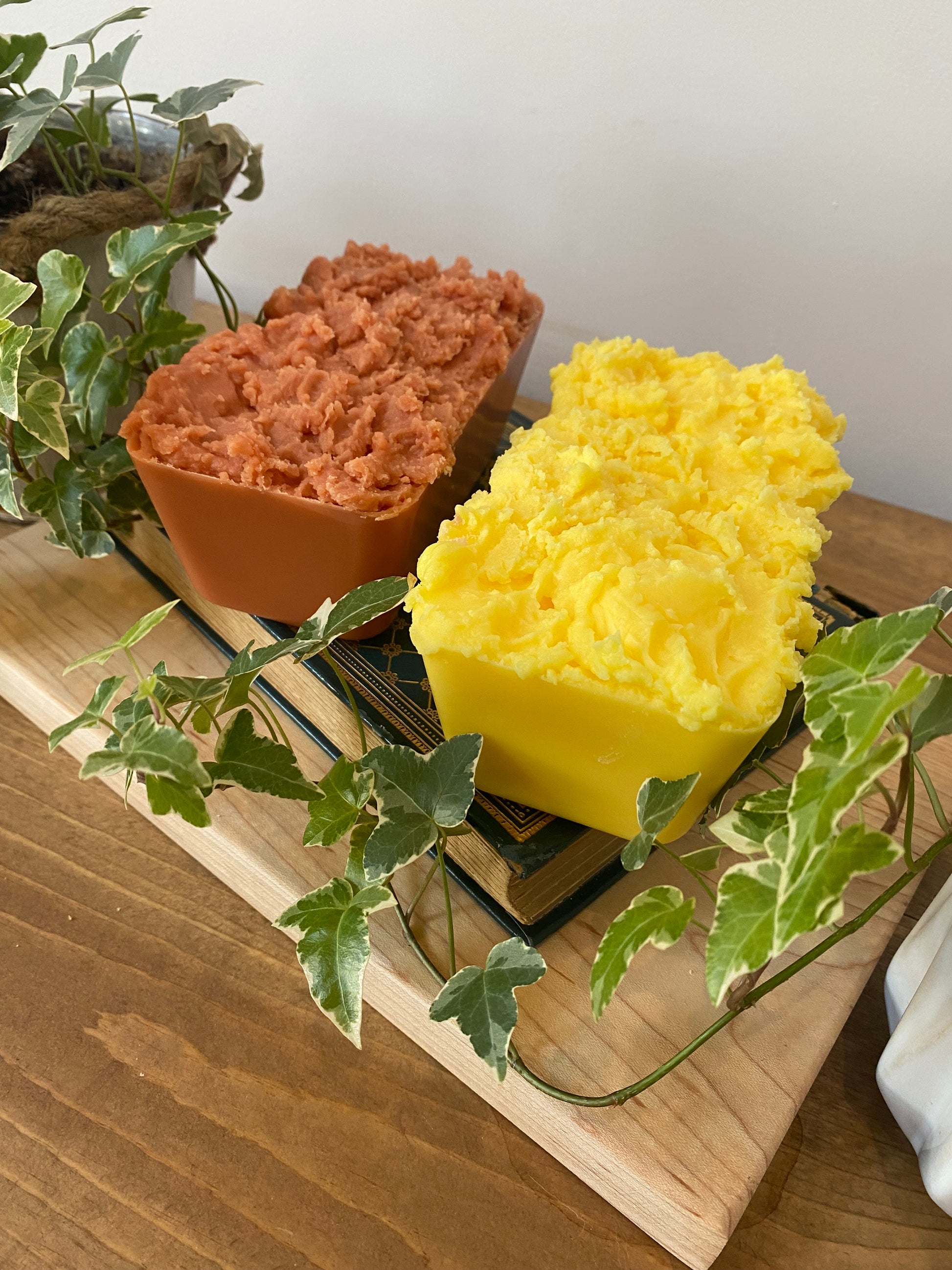 two rectangular objects, one with a yellow, creamy texture and the other with a reddish-brown texture, placed on a wooden surface. There are also some green leaves visible in the background.