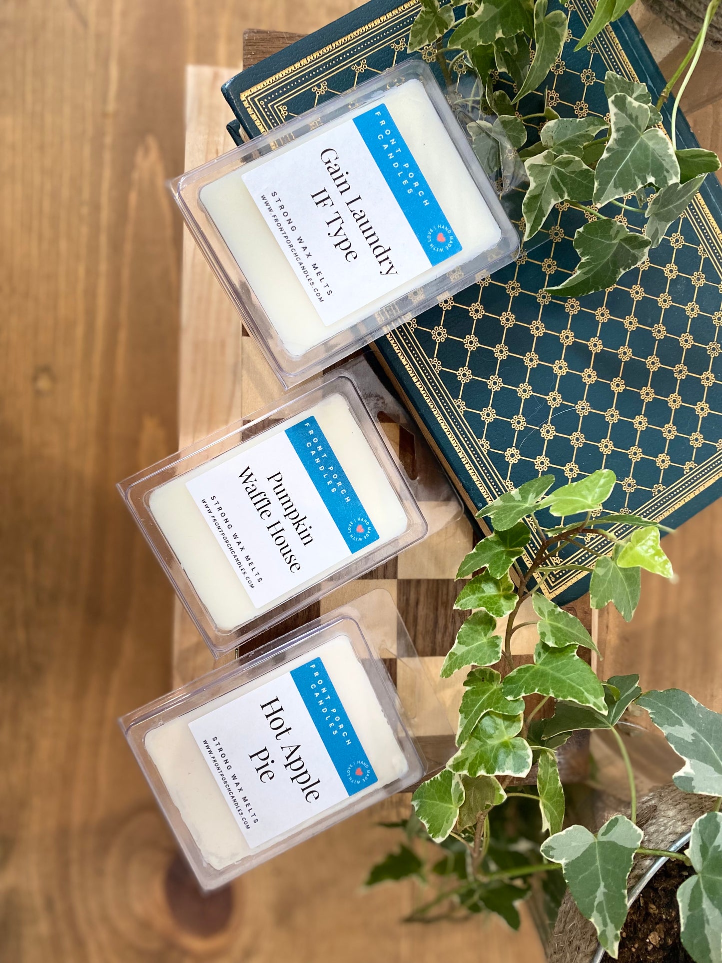 three white candles with blue labels, placed on a wooden surface next to a plant and a book.