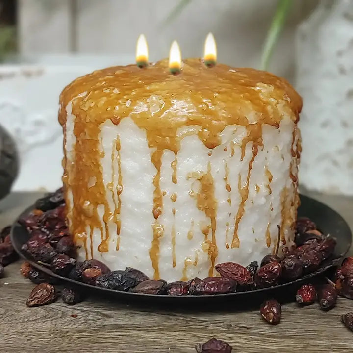 A white cake with caramel drizzle and three lit candles on top, placed on a black plate with dried cranberries.