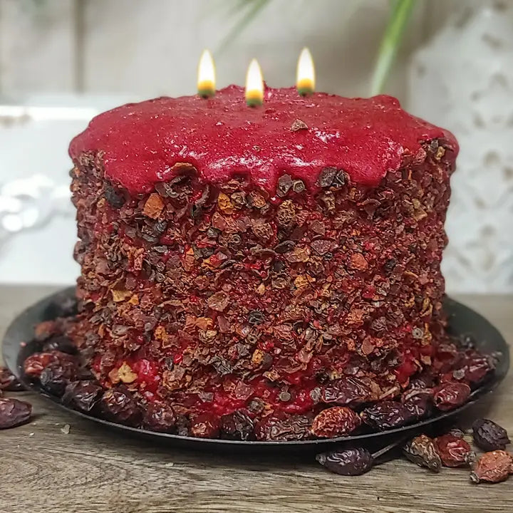 A large, red cake with a red glaze and three lit candles on top, placed on a black plate with dried cranberries around it.