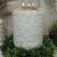 A white, textured candle with three lit candles sits on a wreath of green foliage, with a white vase and potted plant in the background.