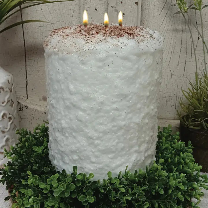 A white, textured candle with three lit candles sits on a wreath of green foliage, with a white vase and potted plant in the background.